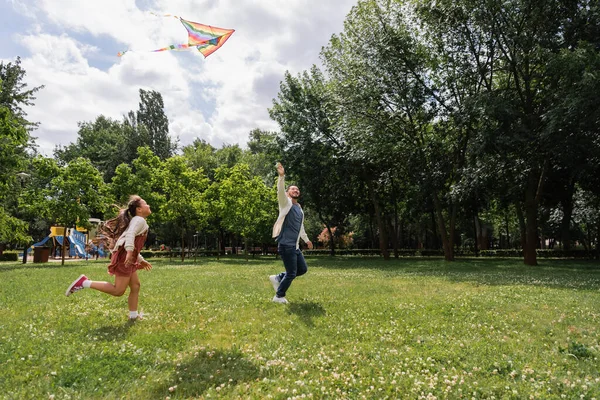 Asian father holding flying kite while running with daughter in park — Stock Photo