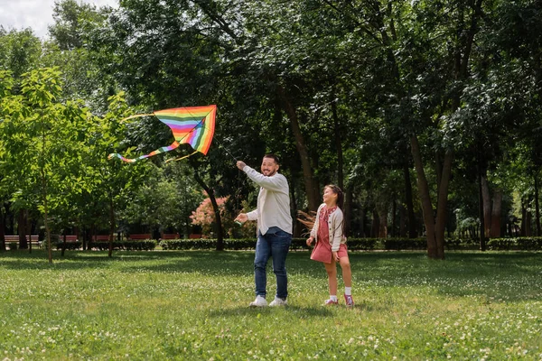 Улыбающийся азиат держит цветного летящего змея рядом с дочерью в парке — стоковое фото