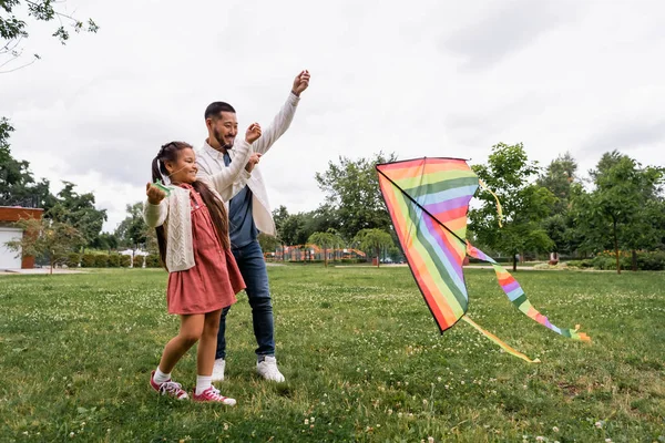 Smiling asian parent and kid looking at flying kite on lawn in park - foto de stock