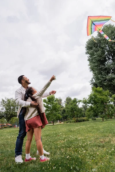 Side view of smiling asian father hugging daughter with flying kite in park - foto de stock