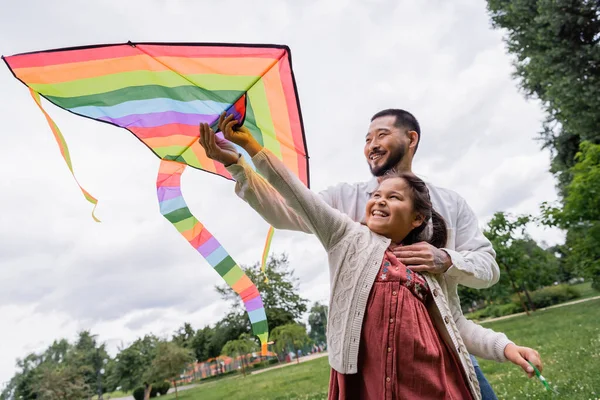 Positive asian dad hugging daughter with flying kite in summer park - foto de stock