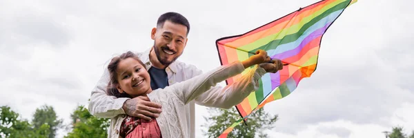 Smiling asian man hugging preteen daughter with flying kite in park, banner - foto de stock