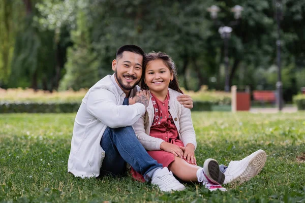 Positive asian man hugging daughter while sitting on grass in summer park — Fotografia de Stock