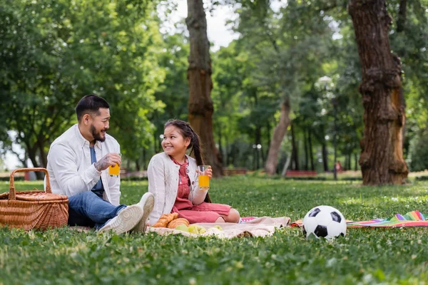 Asian family holding orange juice near food and football on grass in park — Photo de stock