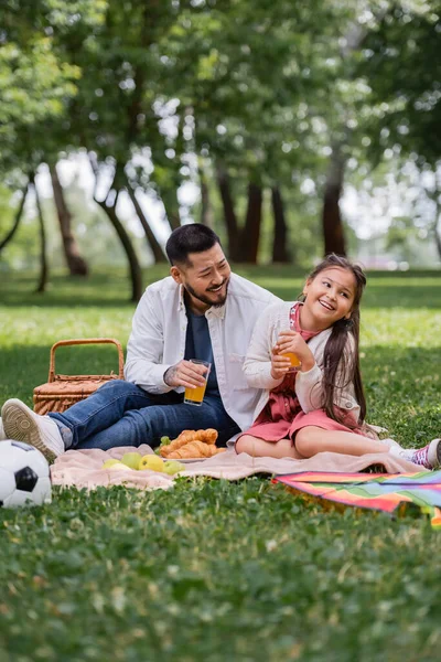 Happy asian family holding orange juice near food and football in summer park - foto de stock