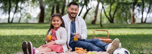 Positive asian child holding orange juice near dad and food in summer park, banner — Photo de stock