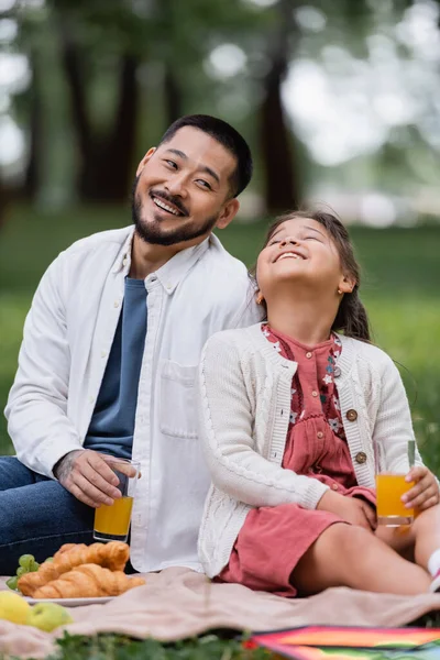 Positive asian family holding orange juice near croissants on blanket in park - foto de stock