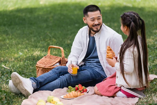Smiling asian parent holding orange juice near daughter with croissant in park - foto de stock