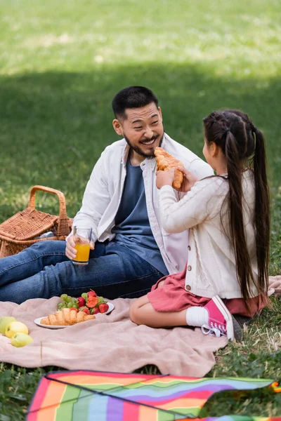 Girl holding croissant near cheerful asian dad with orange juice on blanket in park - foto de stock