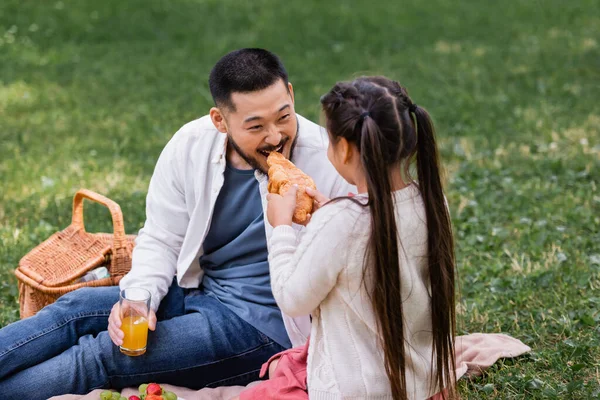 Child holding croissant near asian dad with orange juice in park — Stock Photo