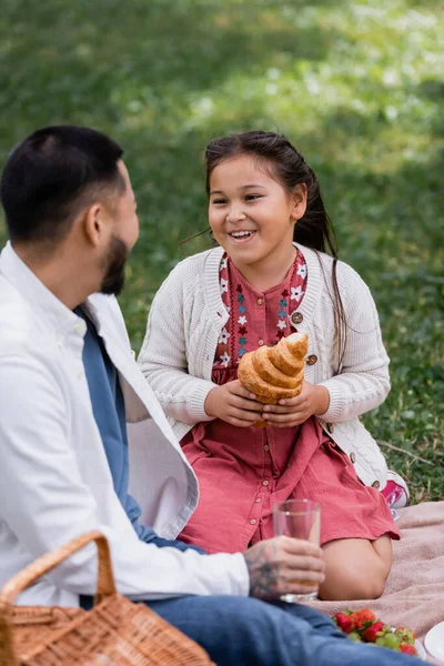 Asian child holding croissant near blurred father holding glass in summer park - foto de stock