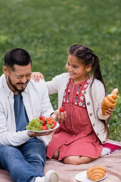 Smiling asian girl holding croissant near cheerful father with fruits in park — Stock Photo