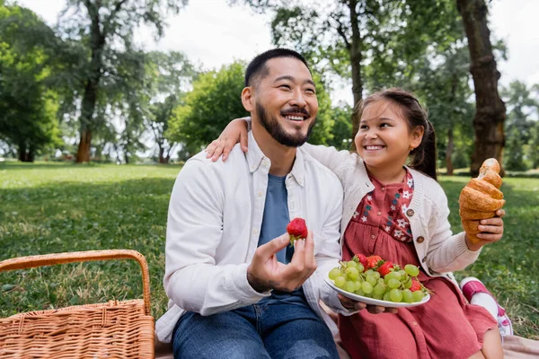 Positive asian girl holding croissant while hugging father with fruits in park — Stock Photo