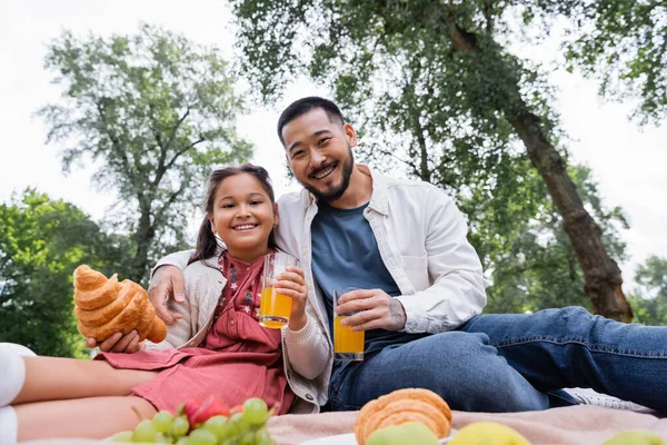 Low angle view of asian family holding orange juice and croissant in summer park — Stock Photo