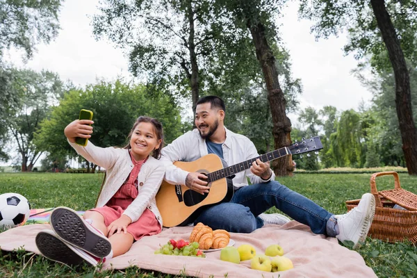 Smiling asian girl taking selfie on smartphone while father playing acoustic guitar during picnic in park - foto de stock