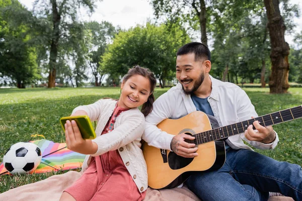 Cheerful asian girl taking selfie with dad playing acoustic guitar near flying kite and football in park - foto de stock