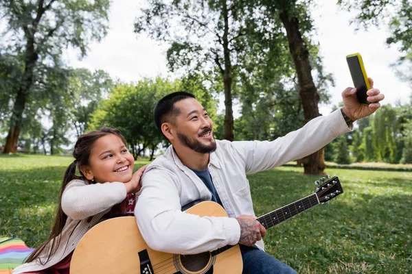 Cheerful asian dad taking selfie on smartphone while holding acoustic guitar near daughter in park — Photo de stock