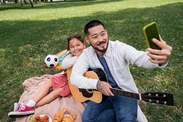 Positive asian man taking selfie on smartphone and holding acoustic guitar near daughter and food in park - foto de stock