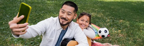 Asian dad taking selfie on cellphone while holding acoustic guitar near child in park, banner — Foto stock