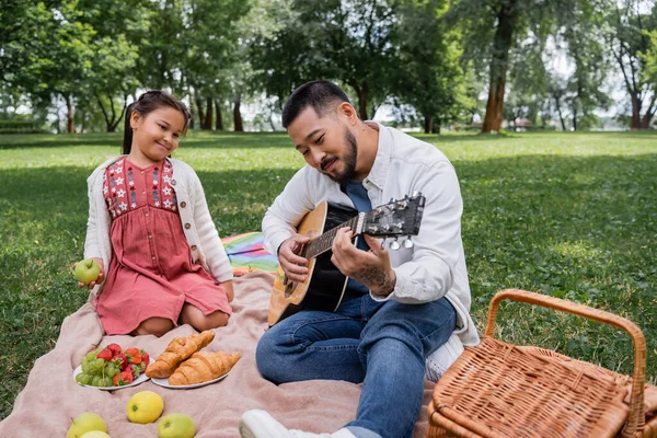 Азіатський чоловік грає на акустичній гітарі біля дочки та їжі в парку. — стокове фото