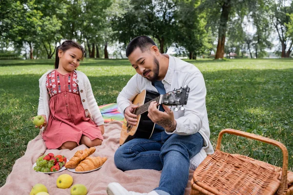 Asian man playing acoustic guitar near daughter and food in park - foto de stock