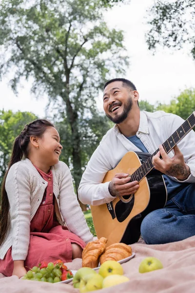 Positive asian parent playing acoustic guitar near daughter and blurred food in park - foto de stock