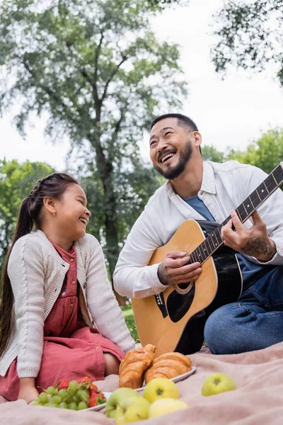 Positive asian parent playing acoustic guitar near daughter and blurred food in park — Stock Photo
