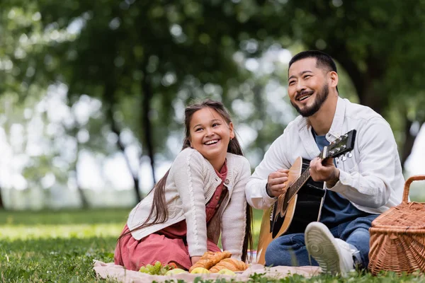Smiling asian dad playing acoustic guitar near daughter and croissants on blanket in park — Foto stock