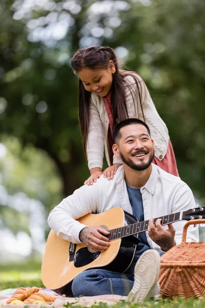 Asian kid standing near dad playing acoustic guitar during picnic in park - foto de stock