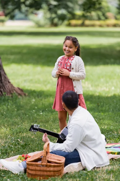 Cheerful asian girl looking at dad playing acoustic guitar during picnic in park — Stockfoto