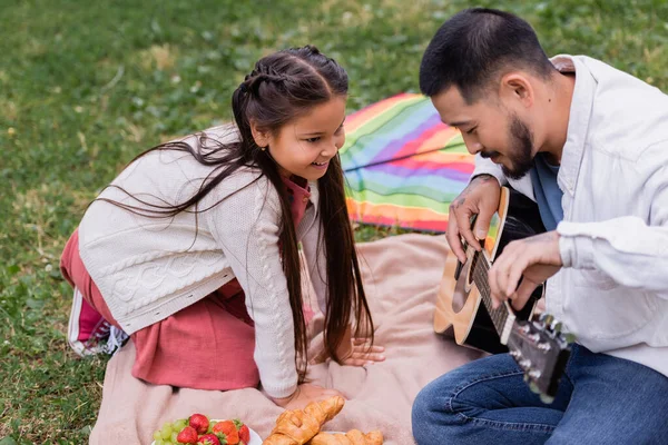Весела азіатська дівчина дивиться на батька, грає на акустичній гітарі біля фруктів і круасанів на ковдрі в парку — стокове фото