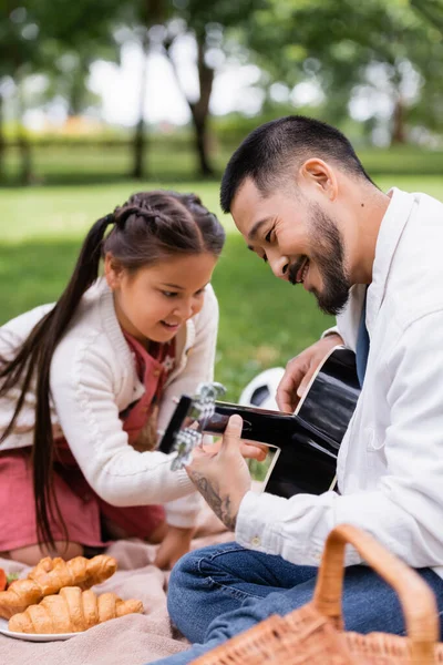 Smiling asian dad playing acoustic guitar near blurred daughter and food in summer park - foto de stock