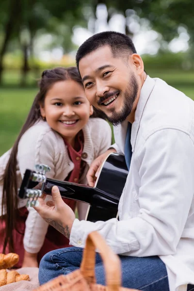 Positive asian man playing acoustic guitar and looking at camera near daughter in park - foto de stock