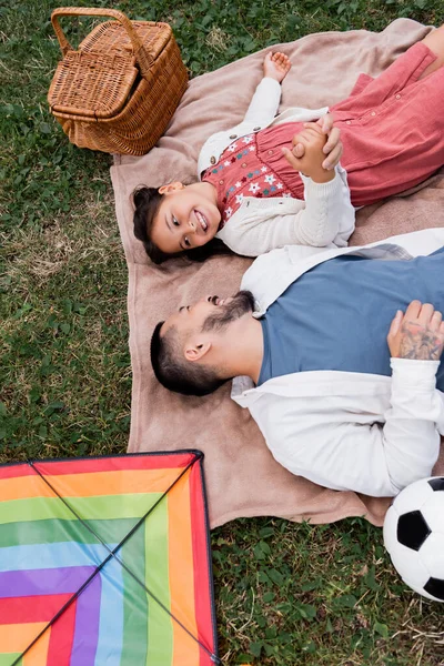 Top view of positive asian father and daughter holding hands while lying on blanket near soccer ball and flying kite in park — Foto stock