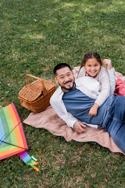 Smiling asian girl hugging father near basket and flying kite in park - foto de stock