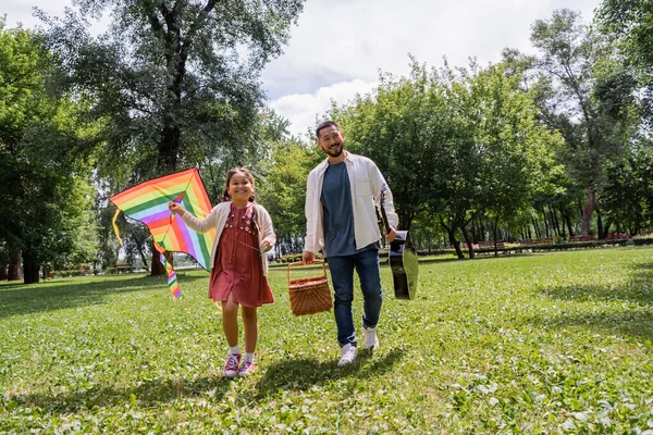 Smiling asian man holding picnic basket and acoustic guitar near daughter with flying kite in park — Foto stock