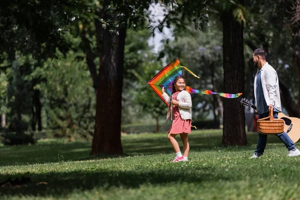 Smiling asian girl holding flying kite near dad with picnic basket and acoustic guitar in park — Stockfoto