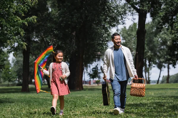 Cheerful asian girl holding flying kite near dad with acoustic guitar and picnic basket in park - foto de stock