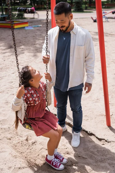 Asian dad looking at daughter on swing in park - foto de stock