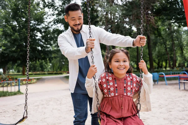 Smiling father standing near asian daughter on swing in park — Photo de stock