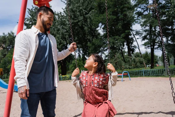Веселий азіатський тато стоїть біля дочки на свінгу в літньому парку — стокове фото