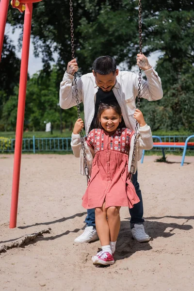 Positive preteen girl sitting on swing near asian dad in park — Stockfoto