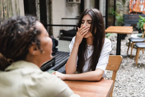 Cheerful woman laughing near blurred african american friend on cafe terrace — Foto stock