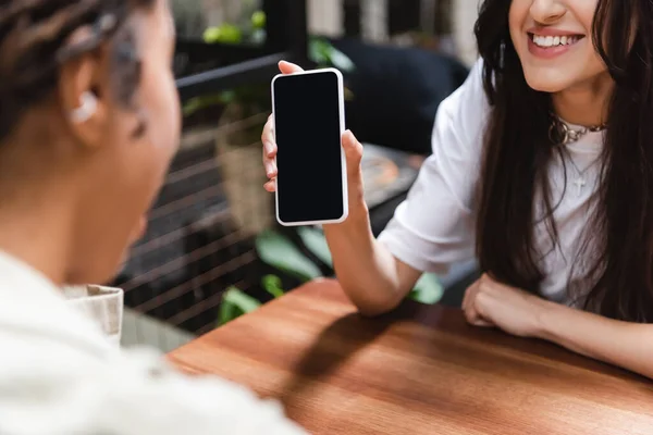 Smiling woman holding smartphone near blurred african american girlfriend in cafe — Stock Photo