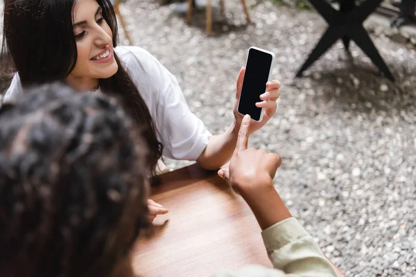 African american woman pointing with finger near friend with smartphone on cafe terrace - foto de stock