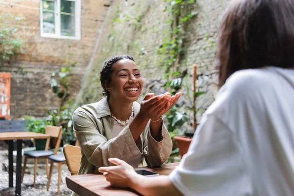 Cheerful african american woman talking to friend near smartphone in outdoor cafe — Foto stock