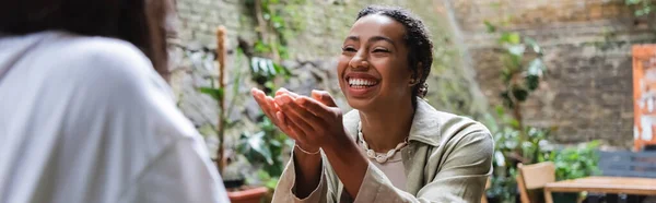 Позитивная африканская американка разговаривает с размытым другом в кафе на открытом воздухе, баннер — стоковое фото