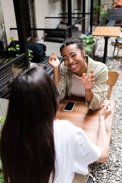 African american woman talking to blurred girlfriend near cellphone in outdoor cafe — Stock Photo