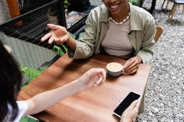 Обрезанный вид на африканскую американскую женщину разговаривает с другом со смартфоном возле кофе в открытом кафе — стоковое фото