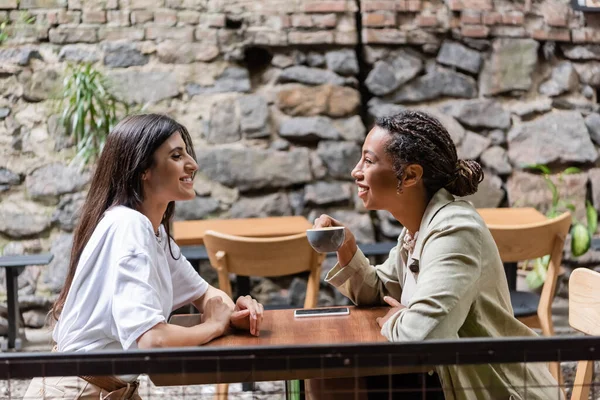 Side view of smiling woman looking at african american friend with coffee on terrace — Stock Photo